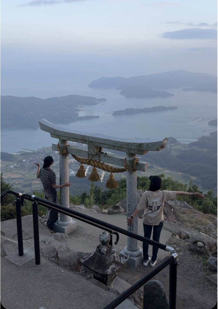 海を見下ろす山頂の鳥居の前で手を広げる様子